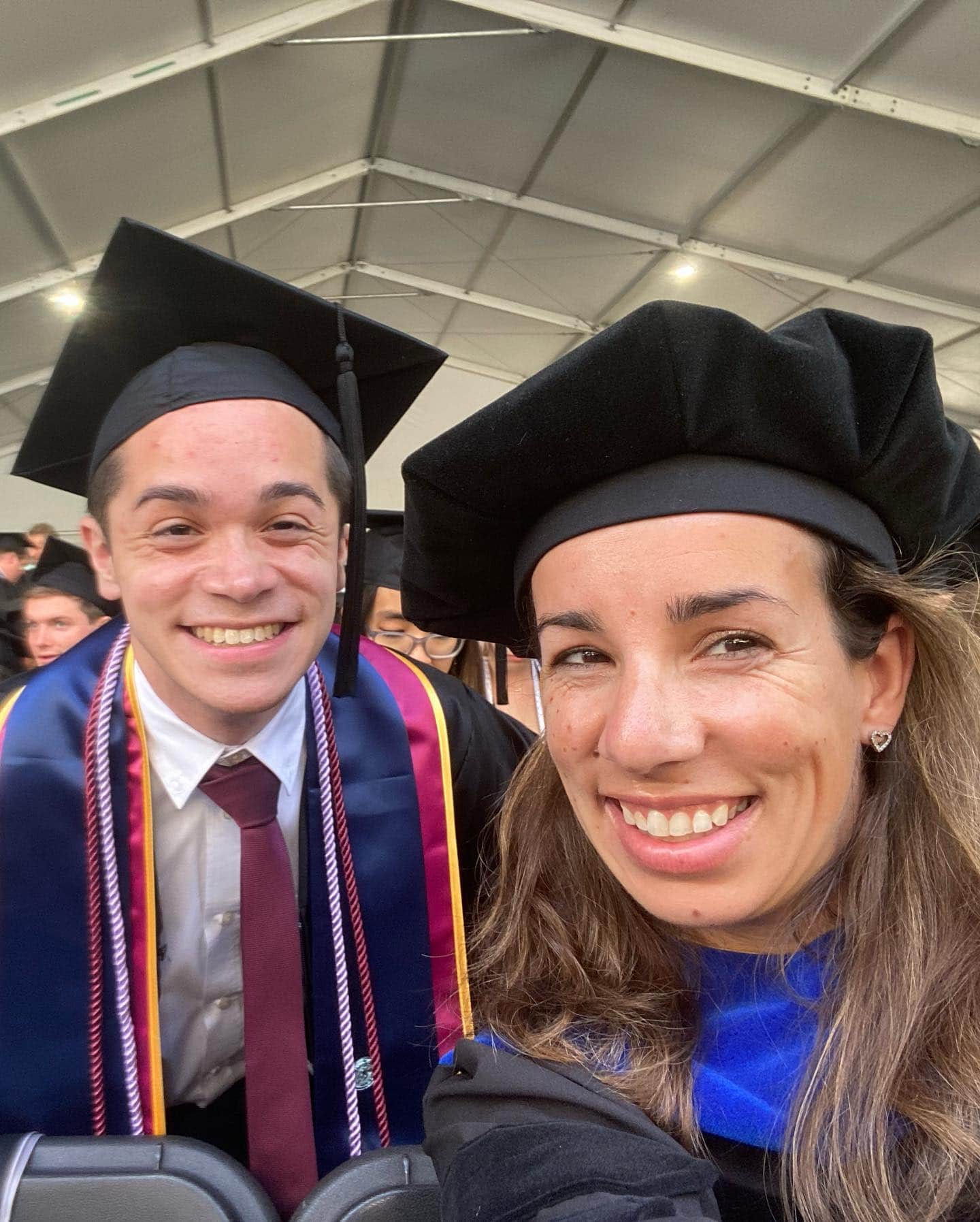A young white man and a slightly older white woman, both wearing academic dress, smile for the camera at graduation.