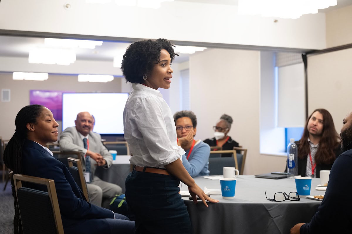 A woman stands before a group, delivering a presentation while the audience attentively listens and takes notes.