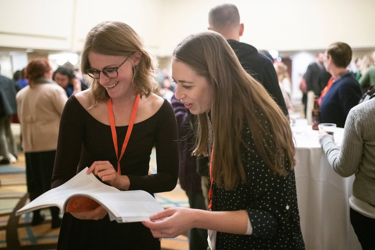 Two smiling annual meeting attendees stand next to each other looking through the same annual meeting program. They stand in a reception room; the background includes attendees mingling at the reception.