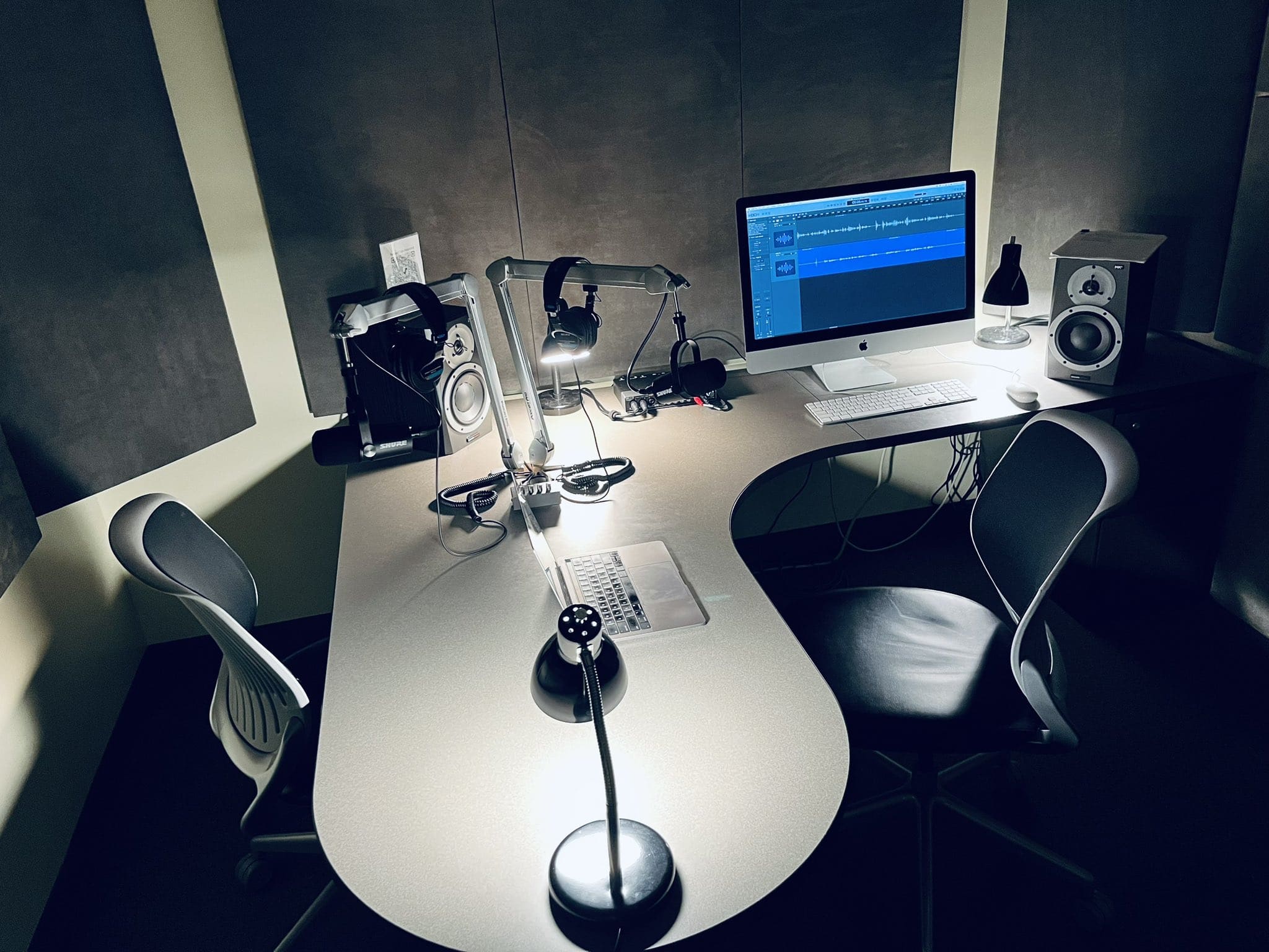 A L-shaped desk with speakers, microphones, headsets, computers, and chairs.