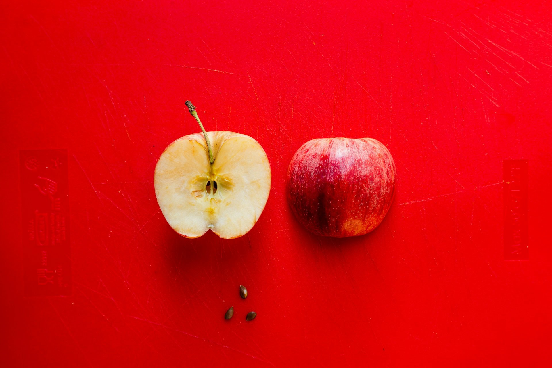 Two halves of a red apple, one face down and one face up, with three seeds below the left half, on a bright red background