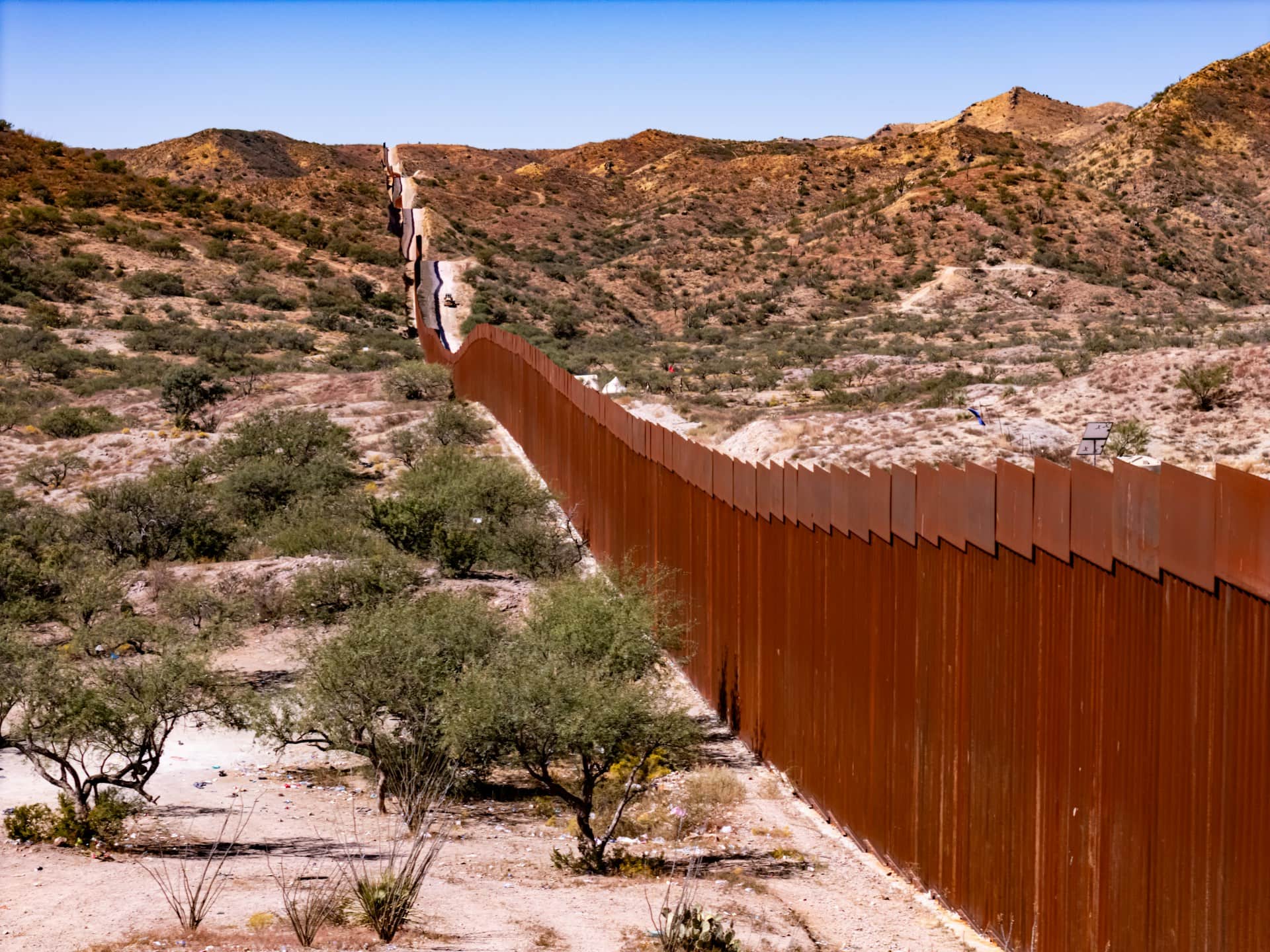 The US-Mexico border wall cutting diagonally across a hilly, arid landscape beneath a sliver of pale blue sky. The wall panels are a rusty brown, while green scrub vegetation dots the white, tan, and brown earth.