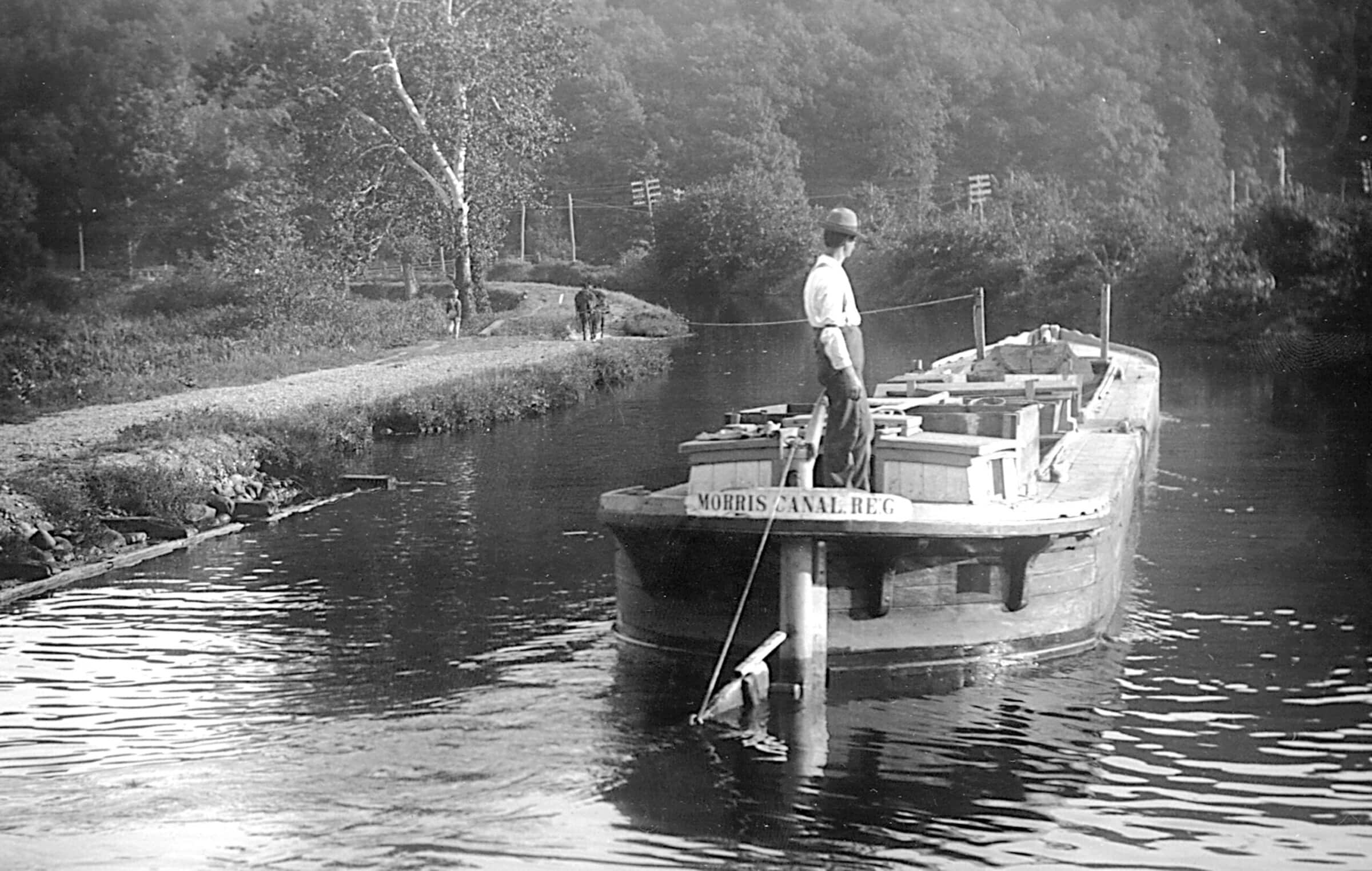 Black and white photograph. A boat on a canal with a man wearing a bowler hat standing at the tiller. A donkey walks ahead of the boat on the canal towpath, connected to the left side of the boat by a rope. The boat has a sign labeling it "Morris Canal Reg." Wooden crates fill the center of the boat.