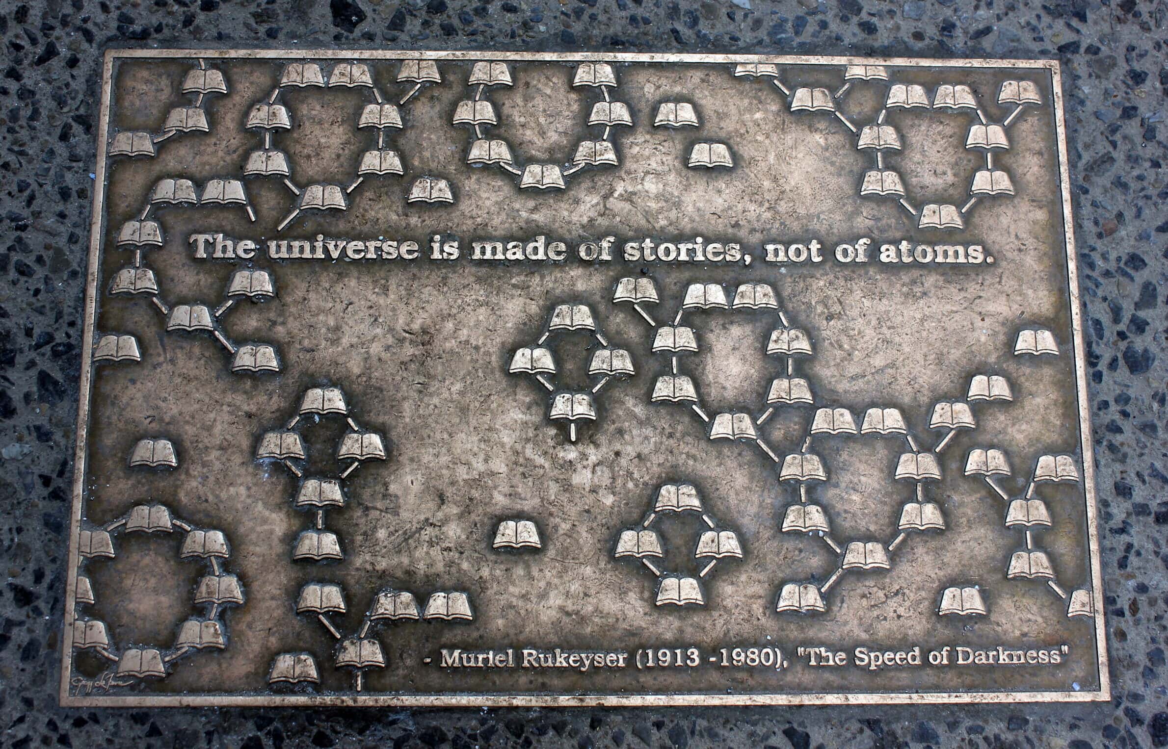 A tarnished bronze plaque, inset into the ground. Books are linked by lines into groupings. It reads "The universe is made of stories, not of atoms." Near the bottom, it reads "-Muriel Rukeyser (1913–1980), "The Speed of Darkness""