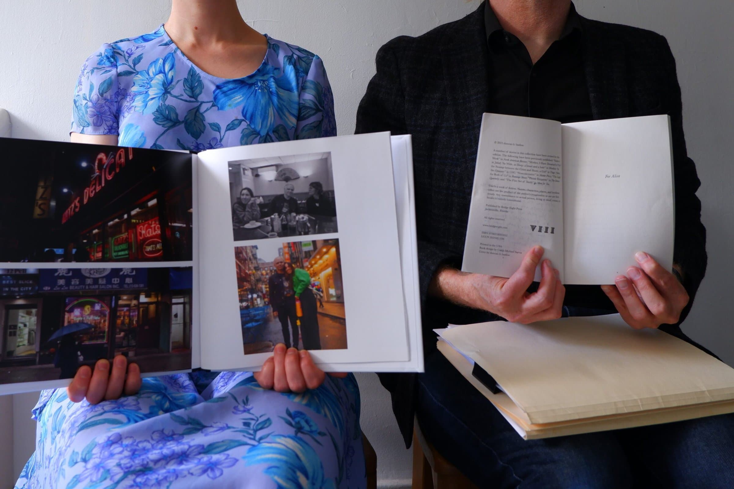 A woman and a man are seated in chairs. The woman holds open a photo book with two photos on each page. The man holds open a book to a dedication page, which reads “For Alisa.” He has manila folders of other papers on his lap.
