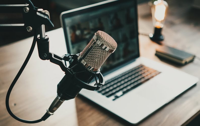 A microphone suspended from a stand outside the image, in front of an open, silver laptop on a wooden table.