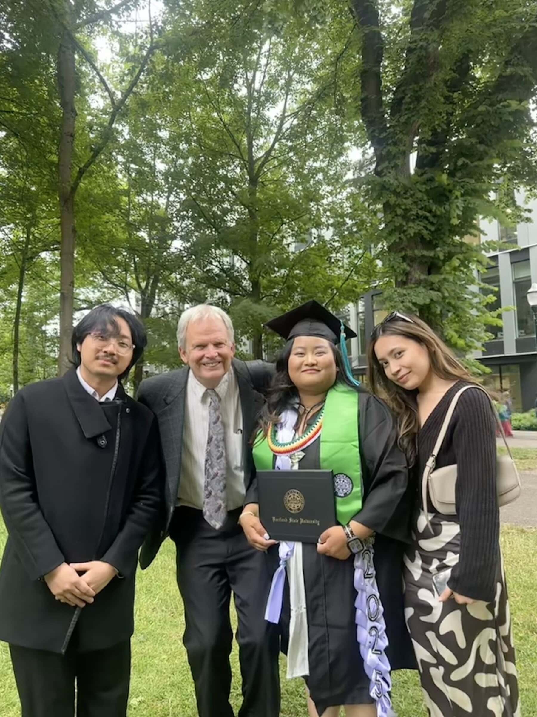 Four people stand in a row in front of trees and a building. The third person is in graduation regalia and holding a diploma.