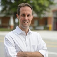 smiling man in a white shirt in front of a tree and building